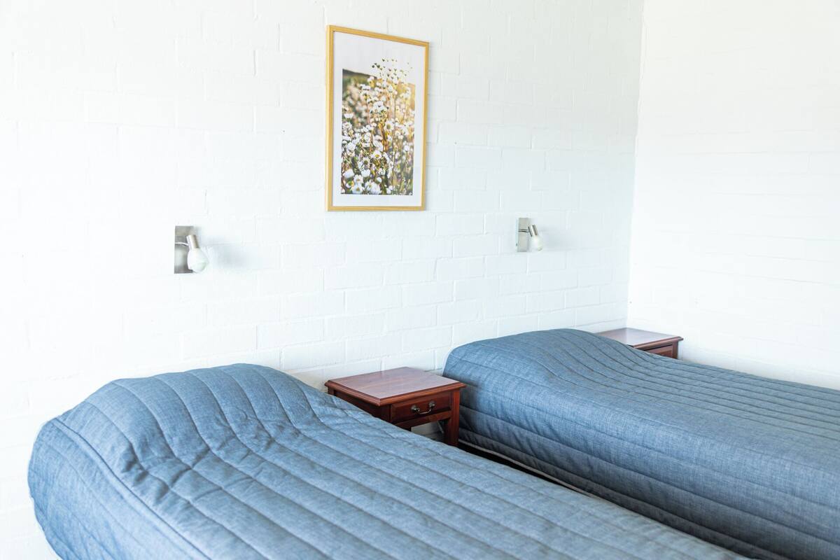 A room with two single beds, a small wooden nightstand, a framed picture on the wall, and two wall-mounted light fixtures.