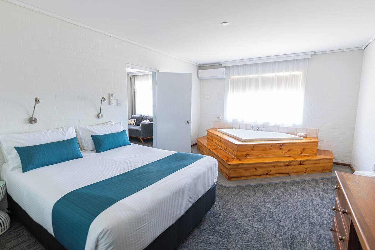 A hotel room with a large bed, a wooden bench with a bathtub, a desk, and a window with curtains.