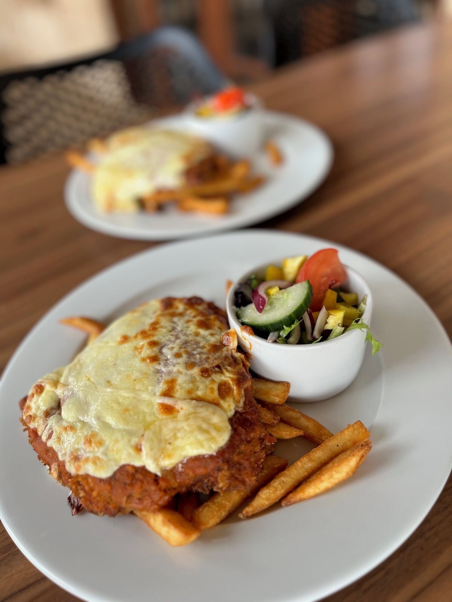 Een bord met een gepaneerde en gebakken schnitzel, overgoten met kaas, vergezeld van frieten en een salade.