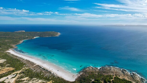 An aerial view of a coastal area with a beach, clear blue waters, and distant landforms.