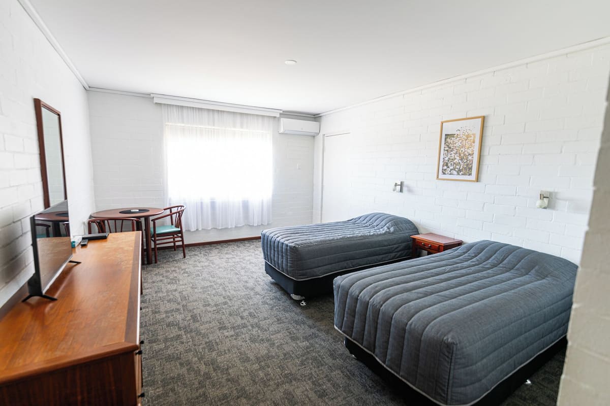 A compact hotel room with two single beds, a small desk, a chair, and a window with curtains.