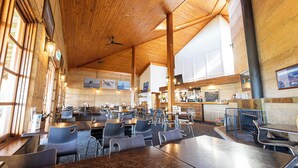 A spacious indoor dining area with wooden ceiling beams, multiple tables and chairs, and large windows allowing natural light.