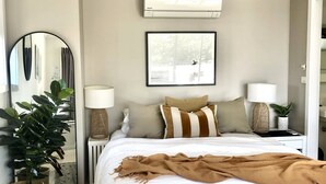 Un dormitorio con una cama, dos mesitas de noche con lámparas, un espejo grande, un cuadro enmarcado en la pared y una planta en maceta.
