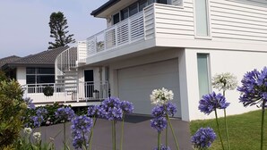 Una casa de dos pisos con balcón, garaje y un jardín bien cuidado con flores moradas.