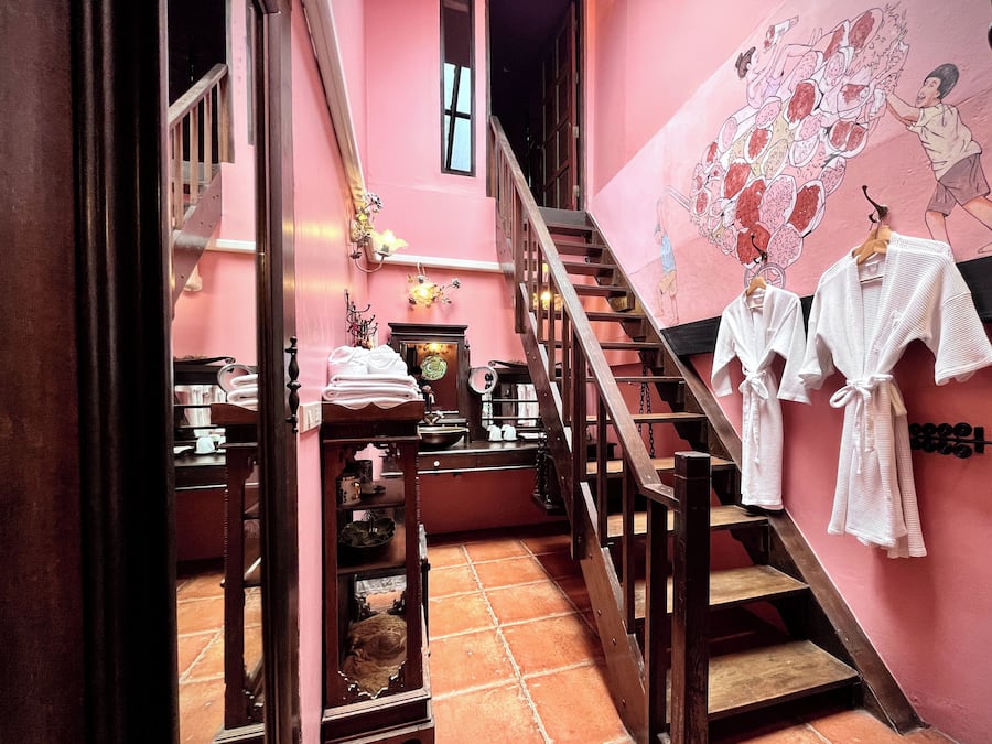 A pink-walled hallway with a staircase, a wooden railing, and a wall-mounted rack with white robes.