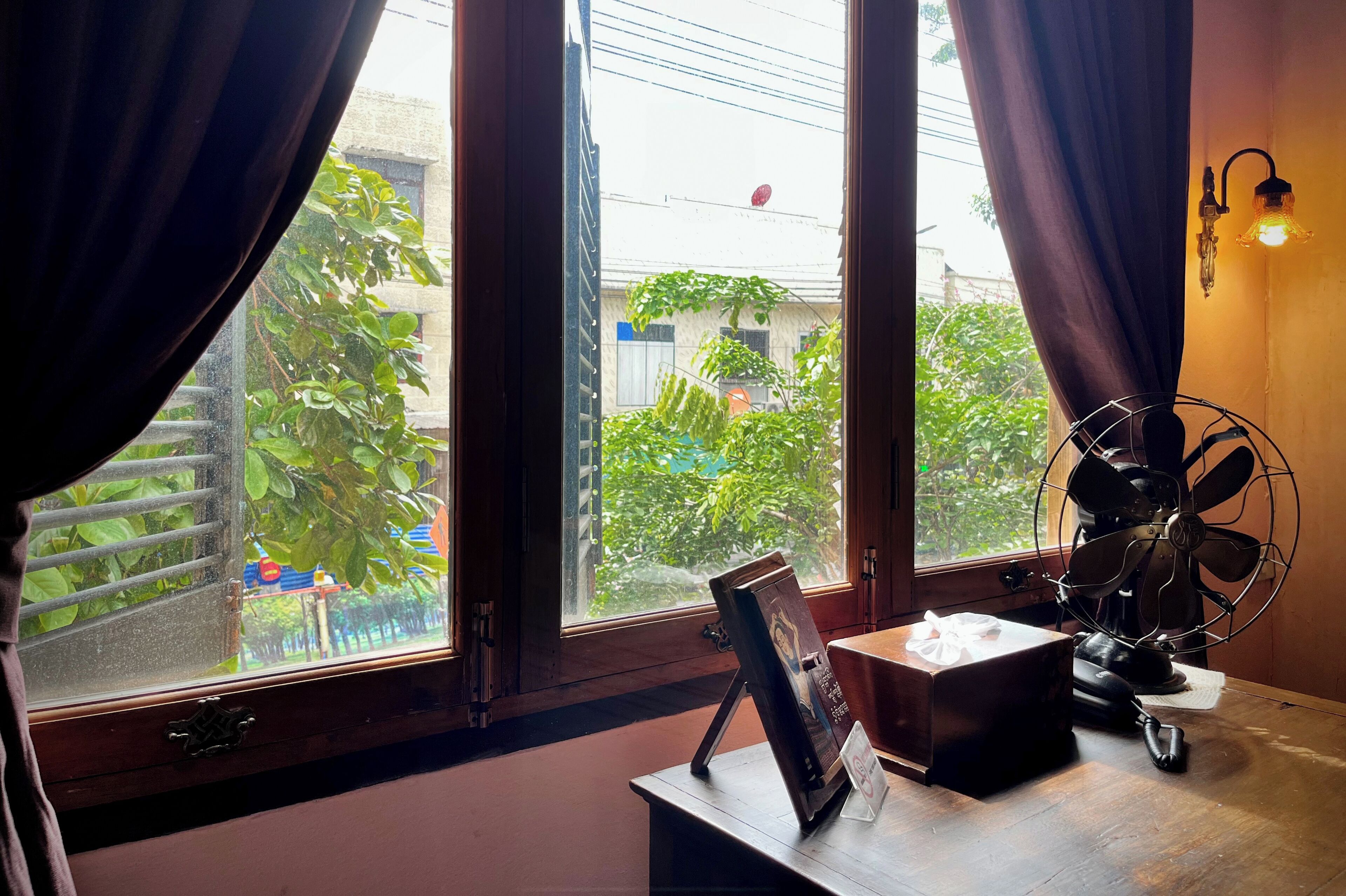 A room with a large window, a wooden desk, a vintage fan, and a picture frame.