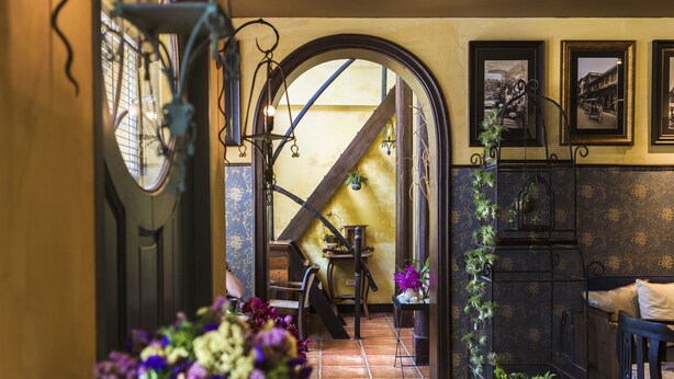 A narrow hallway with a staircase leading to a second floor, a wooden chair, a table, and a plant arrangement. - Old Capital Bike Inn (Bangkok)