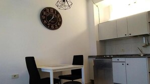 Une petite cuisine avec une table blanche et deux chaises noires, une horloge au mur et des placards blancs.