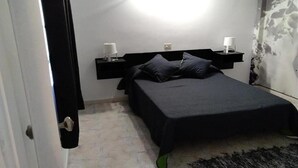 Une chambre à coucher avec un lit, une table de chevet, une lampe fixée au mur et une vue partielle sur une porte.