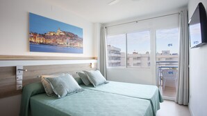 Una habitación de hotel con cama, un cuadro de una ciudad costera, un balcón con una silla y un televisor en la pared.