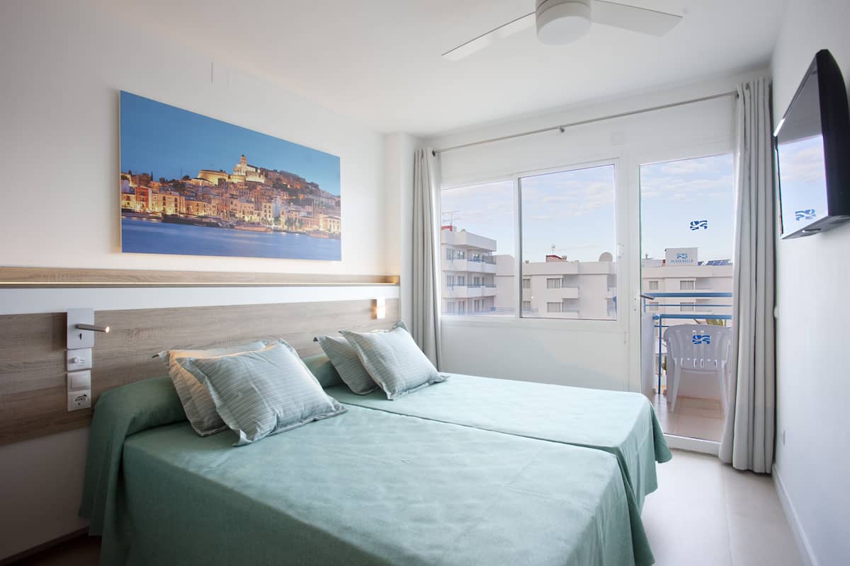 A hotel room with a bed, a painting of a coastal city, a balcony with a chair, and a TV on the wall.