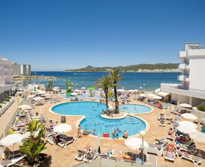 A rooftop pool area with multiple swimming pools, sun loungers, umbrellas, and a view of the sea and distant buildings. - Playa Bella Beach Apartments (Sant Josep de sa Talaia)