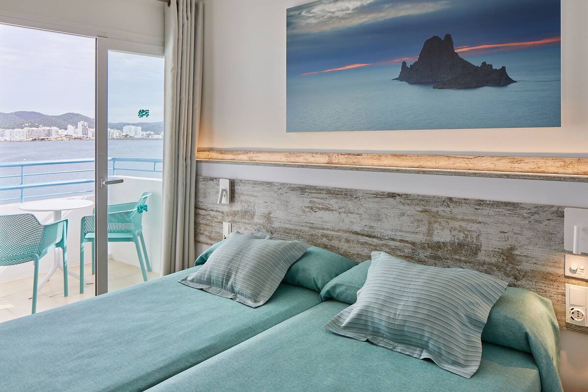 A hotel room with a bed, a view of the sea, and a balcony with chairs.
