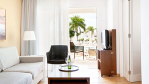 A hotel room with a sofa, a dining table with a tray of wine, a TV, and a view of the outdoor area with palm trees. - Sunprime Atlantic View Suite & Spa (San Bartolomé de Tirajana)