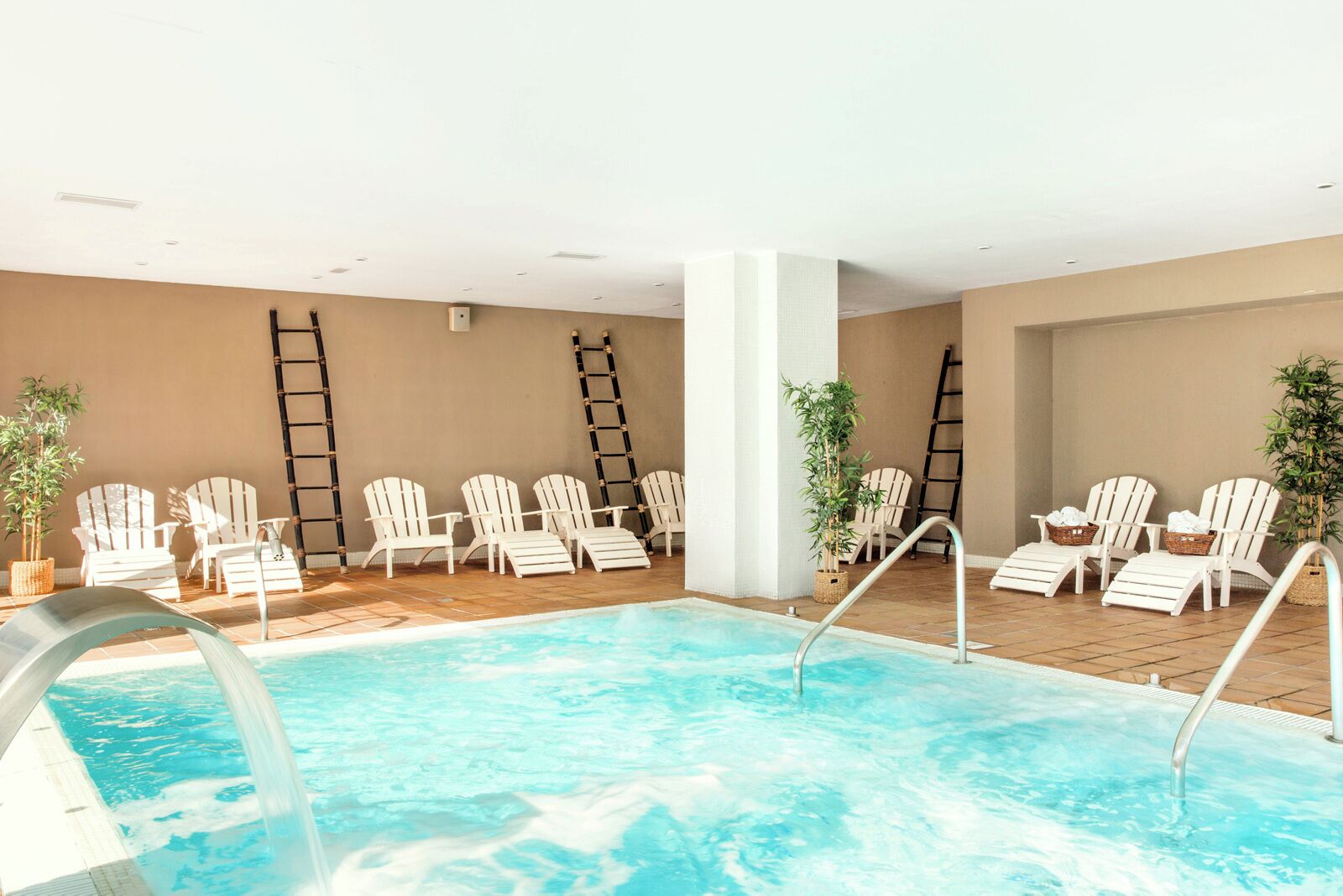 A swimming pool with white lounge chairs and ladders, surrounded by beige walls and potted plants.