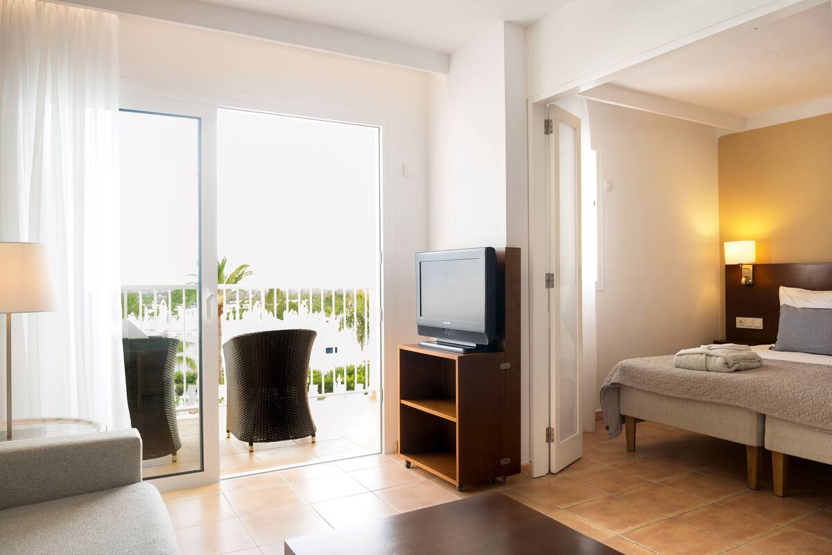 A hotel room with a bed, a TV on a wooden stand, a sofa, a chair, and a balcony with a view.