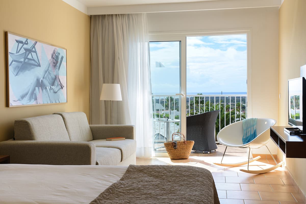 A hotel room with a bed, a sofa, a chair, a TV, and a balcony with a view.