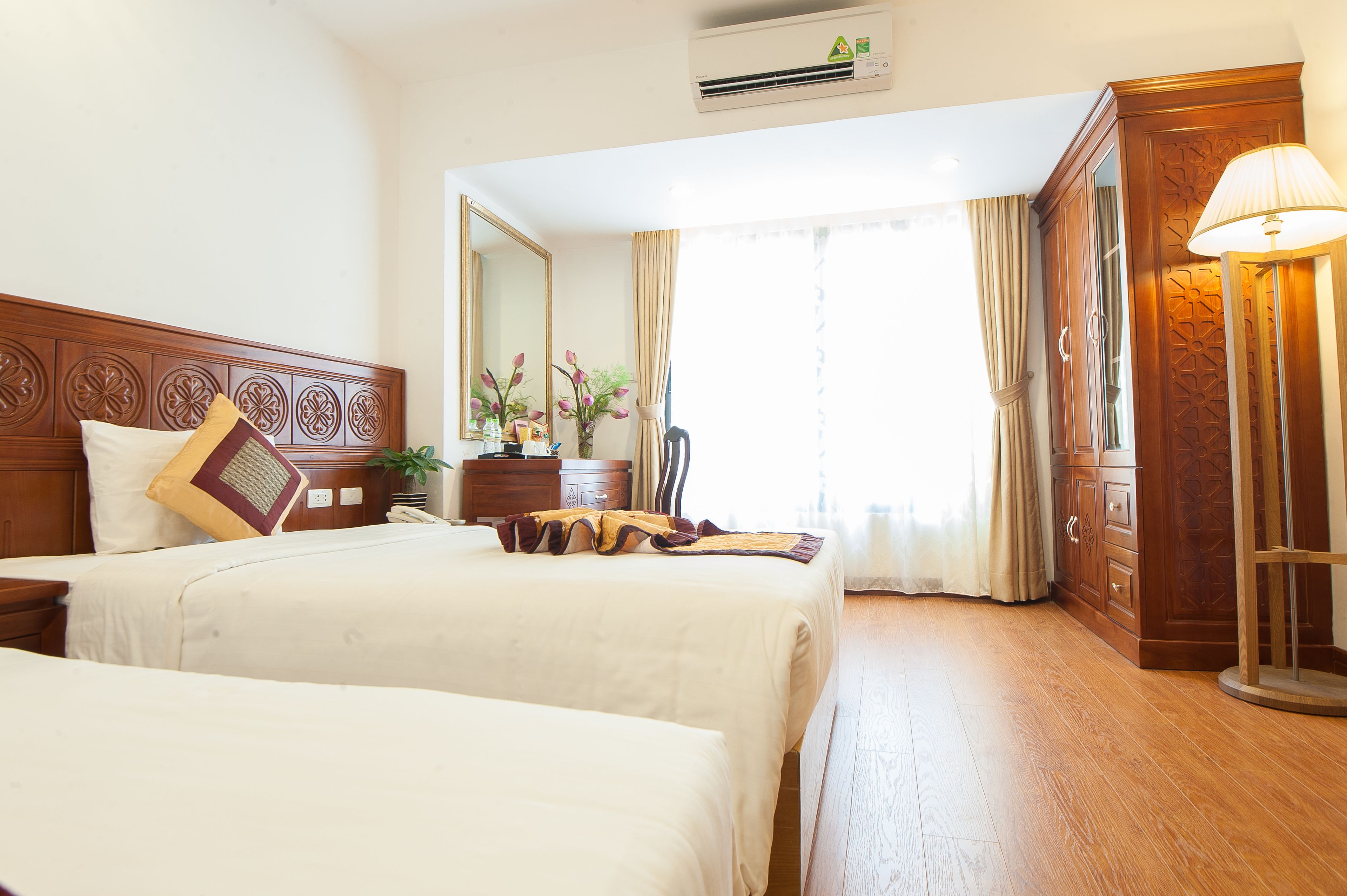 A hotel room with two beds, a wooden headboard, a nightstand, a dresser, a mirror, a lamp, and a window with curtains.