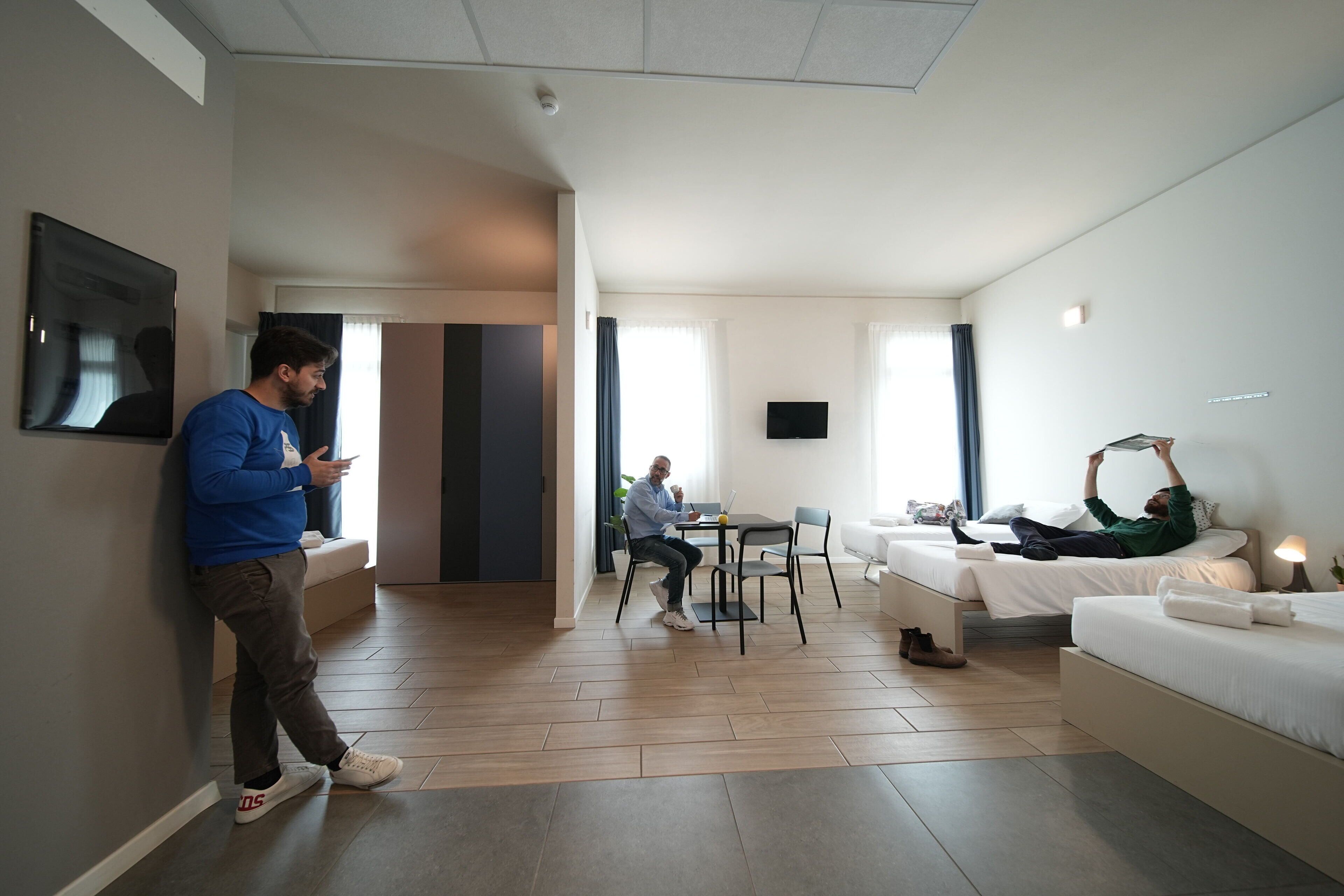 A modern hotel room with a dining area, a TV, and a person lying on the bed.