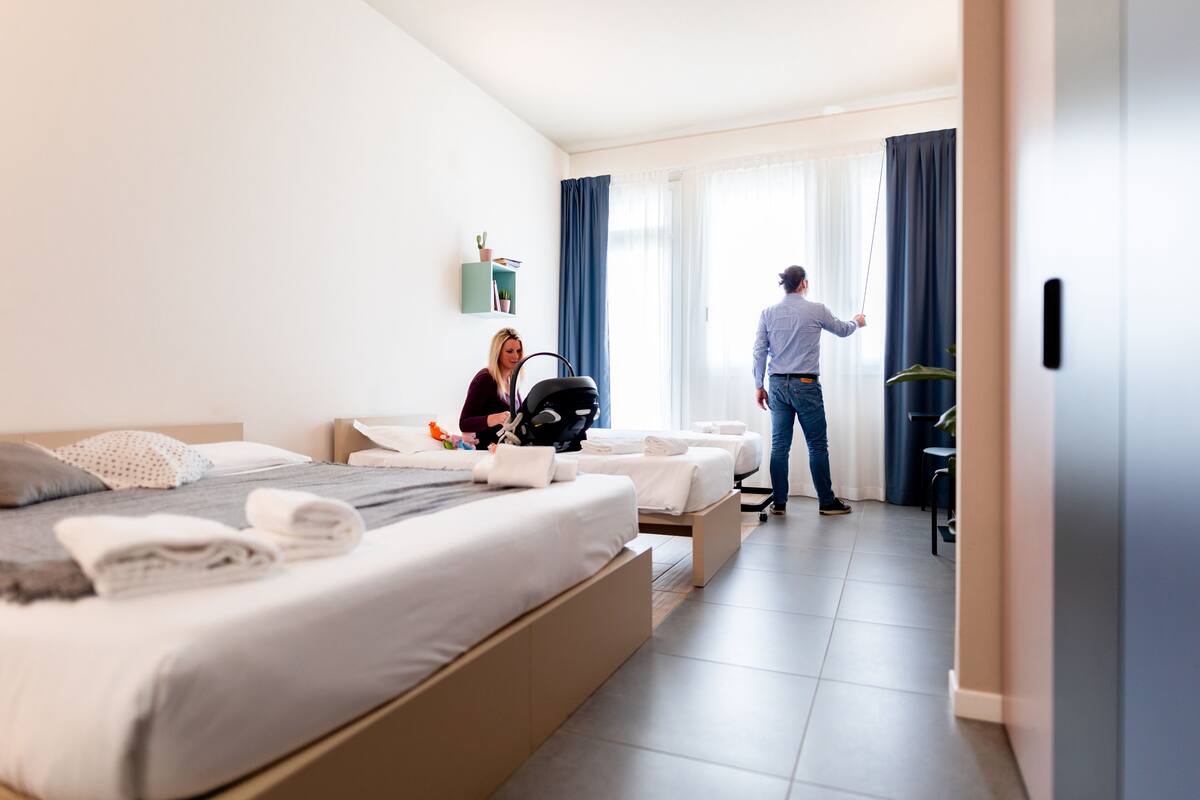 A modern hotel room with a large bed, a desk, and a chair. There is a suitcase on the bed and a person standing near the door.