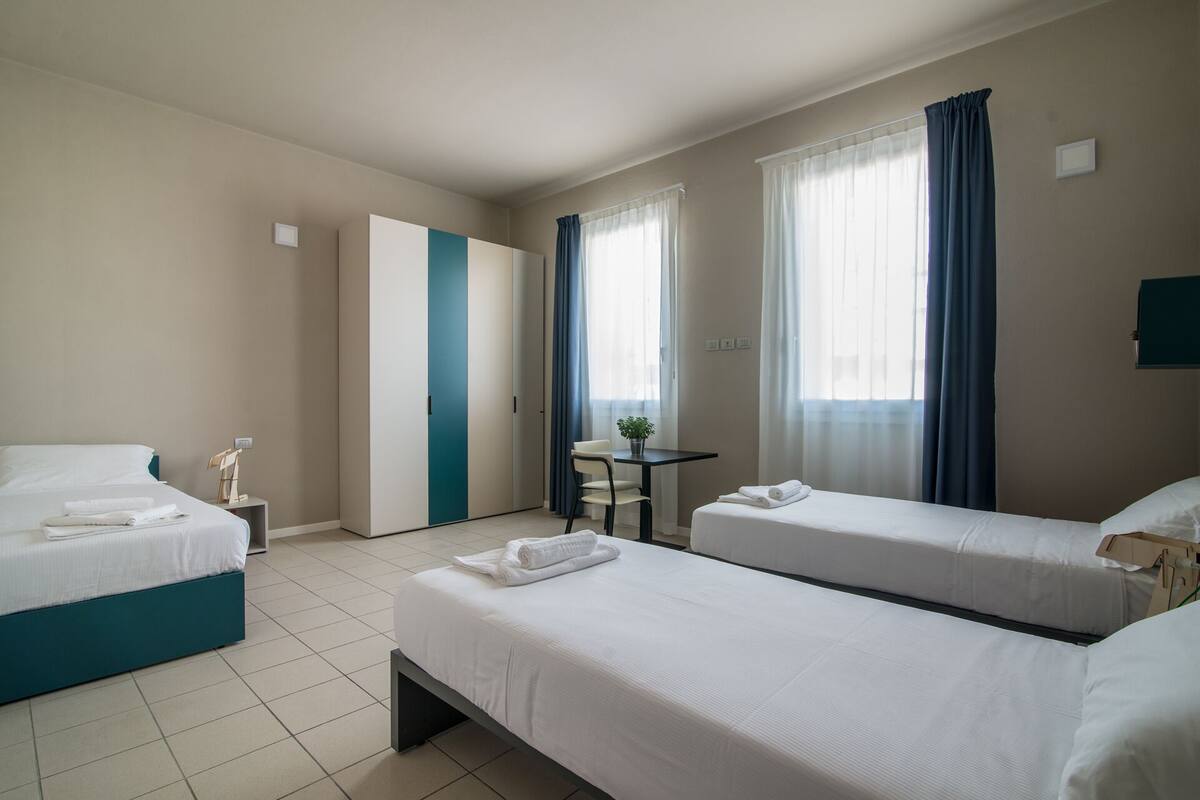 A hotel room with two beds, a desk, and a chair. There is a door and a window with curtains.