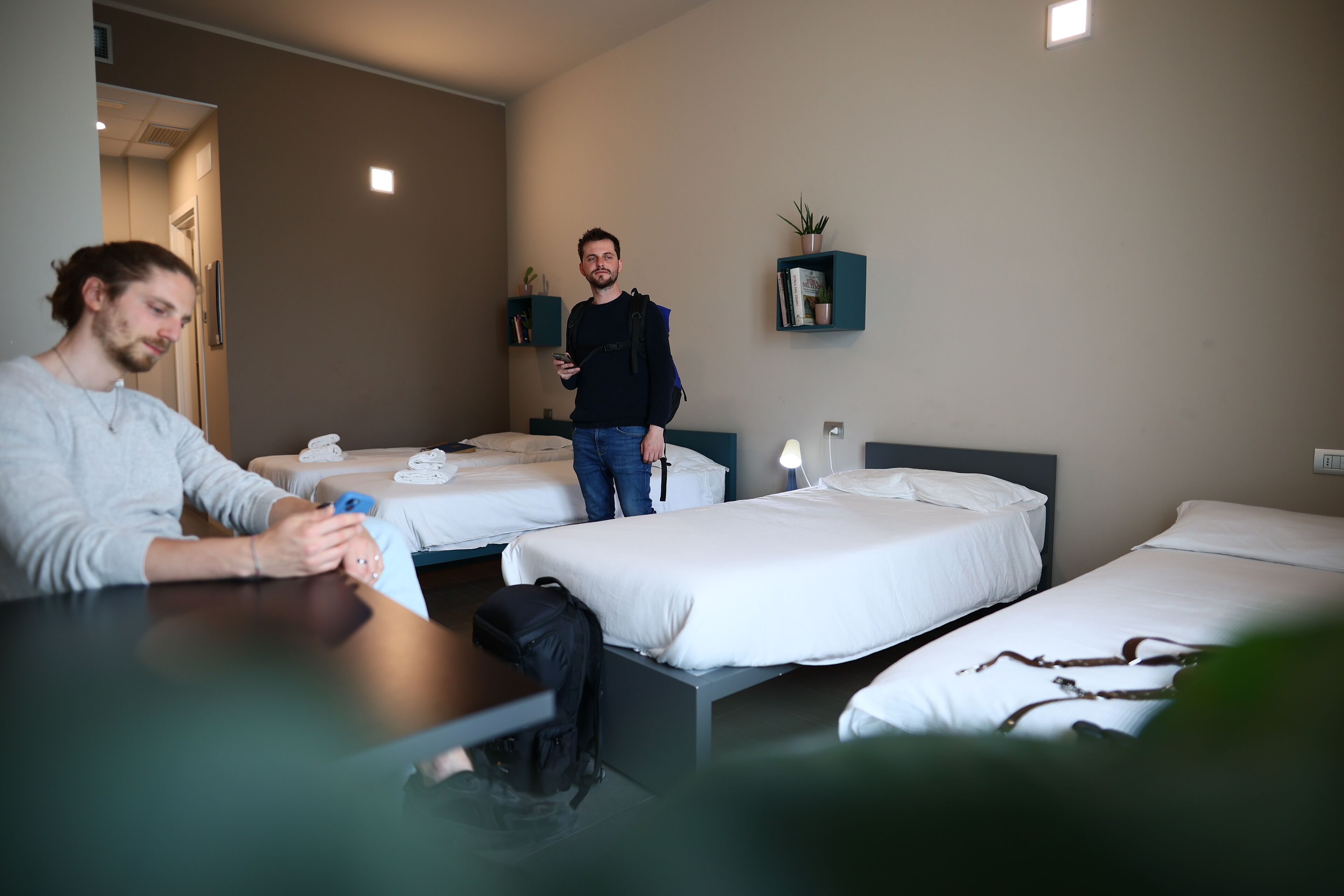 Een hotelkamer met twee eenpersoonsbedden, een tafeltje en een stoel. Iemand zit aan de tafel en gebruikt een smartphone. Een andere persoon staat in de buurt van de deur.