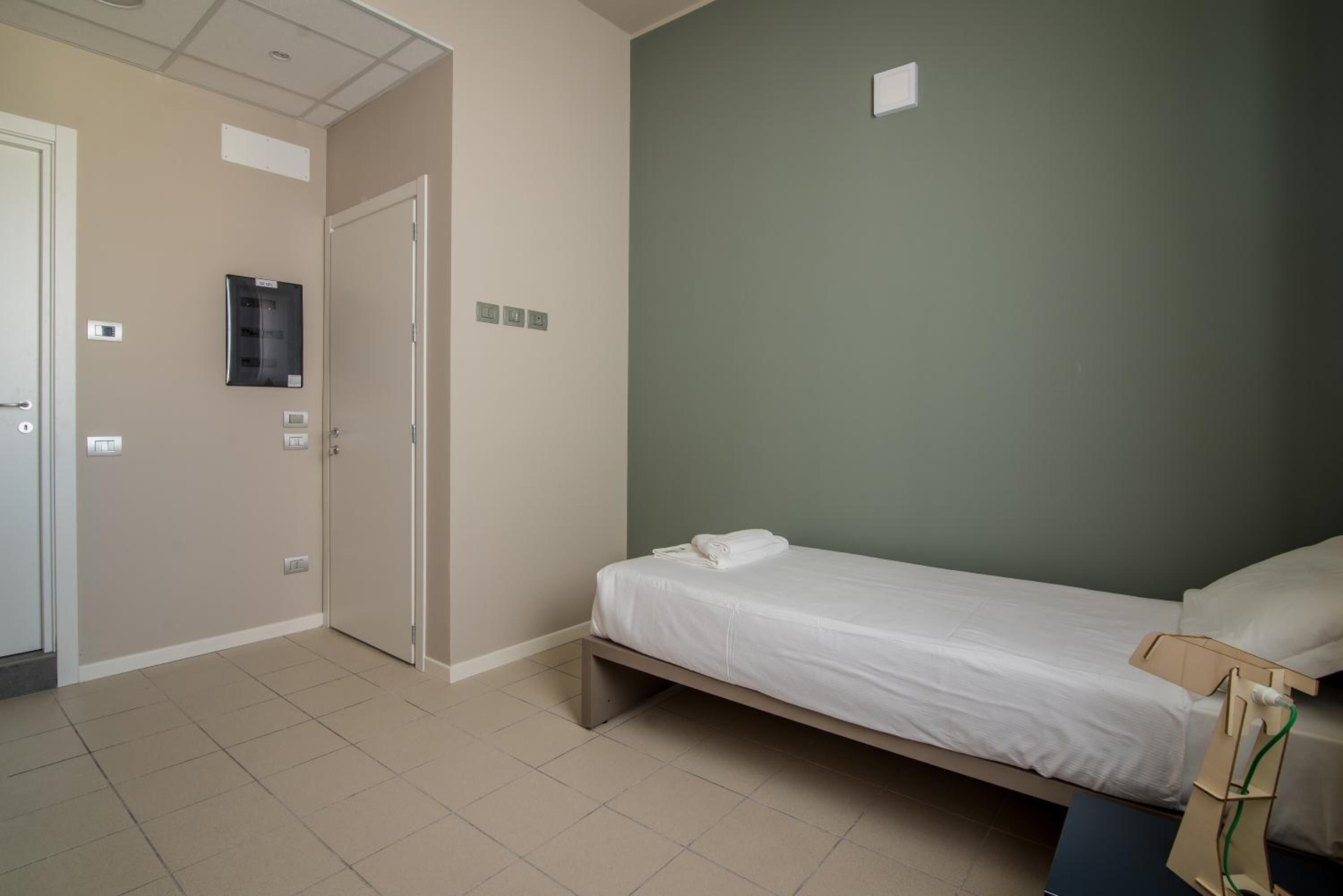 Una habitación pequeña y limpia con una cama individual, una puerta y un panel de control en la pared.