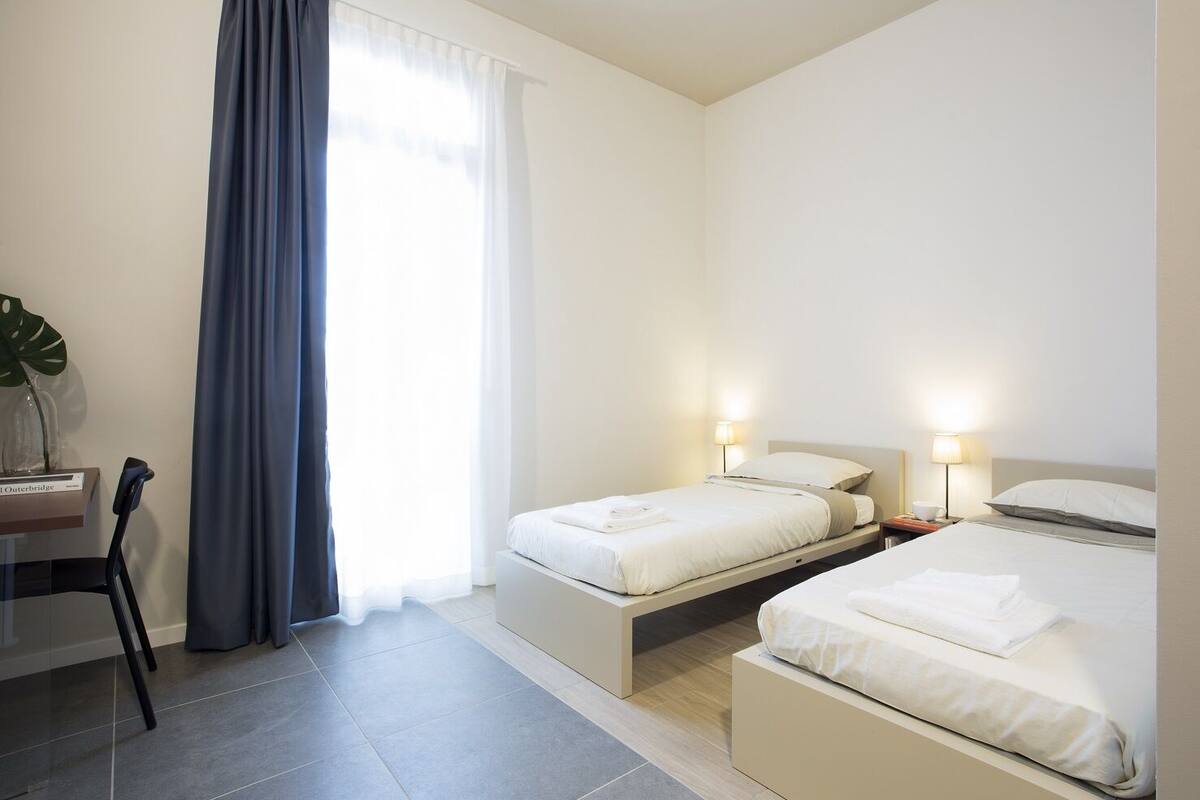 A hotel room with two beds, a desk, a chair, and a large window with curtains.