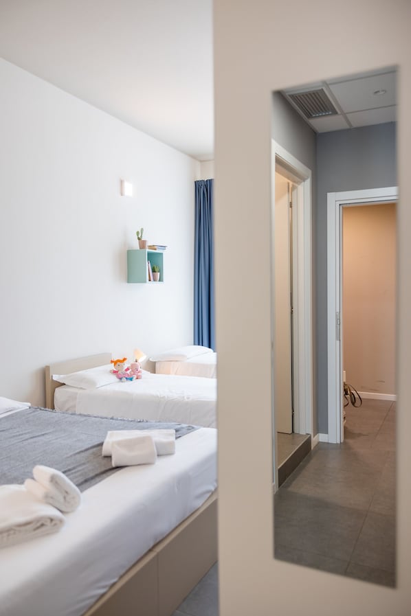 Habitación de hotel con dos camas, un estante pequeño con libros y una planta, y vista al pasillo.