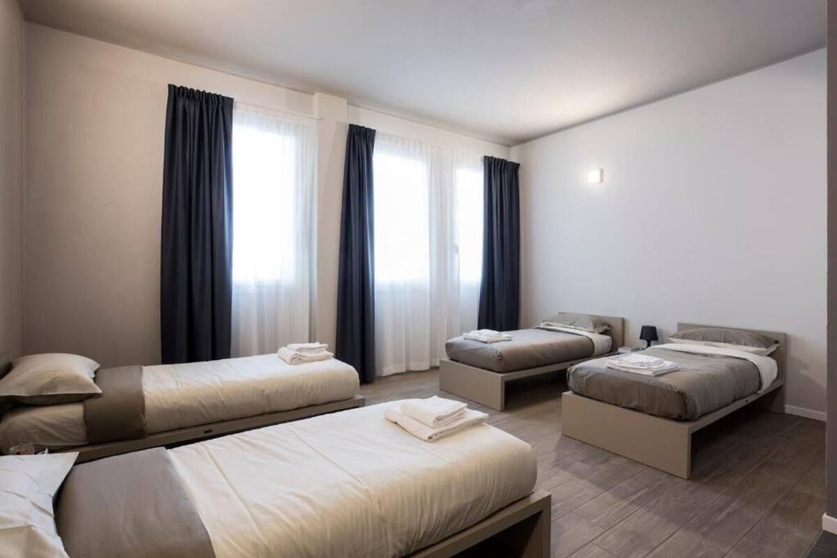 A hotel room with three beds arranged in a row, a large window with curtains, and a bedside table with a lamp.