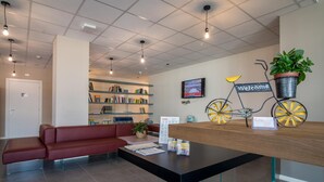 A reception area with a red sofa, a black table with brochures, a 'Welcome' sign on a bicycle, and a television displaying a scenic view. - Casa A Colori Padova (Padova)