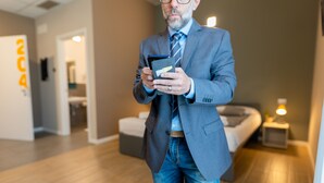 A man in a suit holding a tablet in a modern hotel room with a bed, bedside table, and a small nightstand with a lamp. - Casa A Colori Padova (Padova)