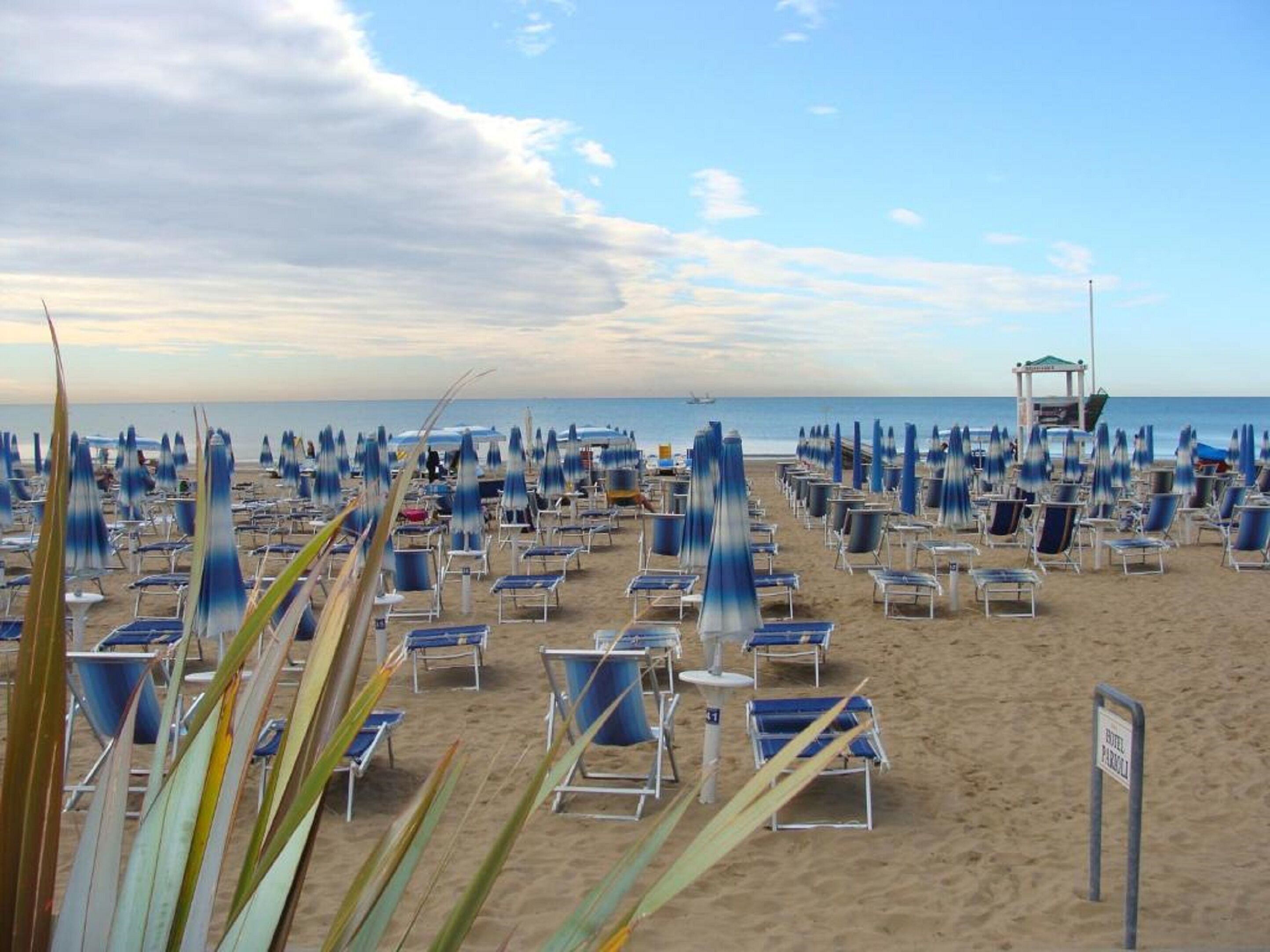 Une plage bordée de rangées de chaises de plage et de parasols bleus et blancs, une tour de surveillance des sauveteurs et un panneau indiquant la présence d’un hôtel.