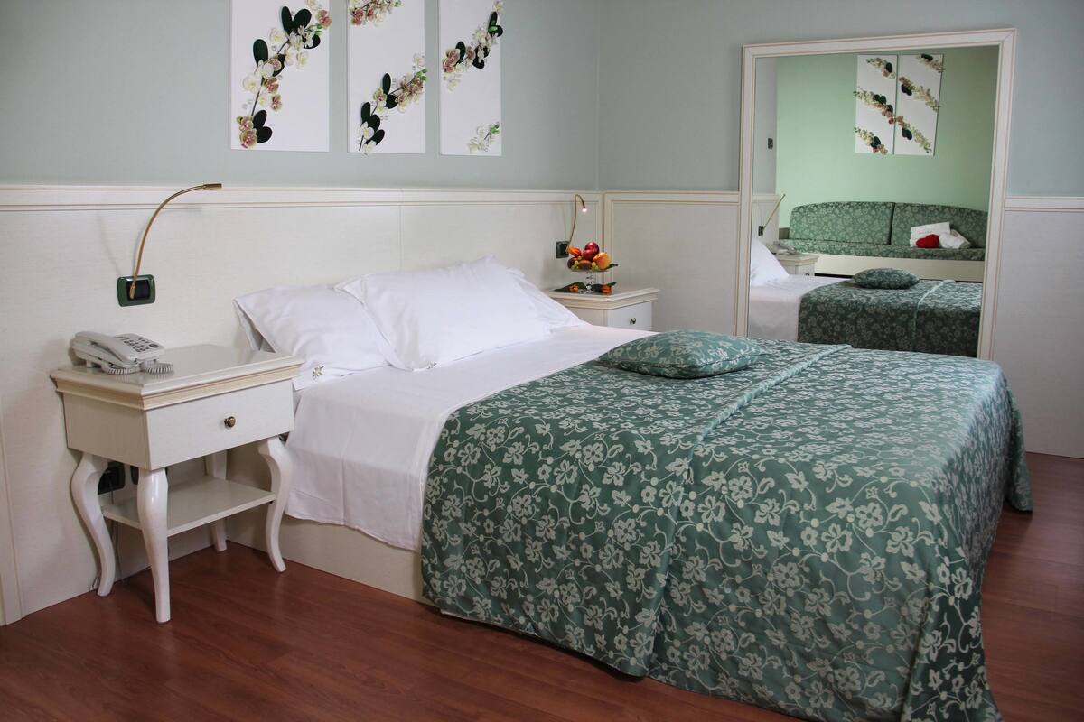 Ein Schlafzimmer mit einem Einzelbett, einem Nachttisch mit Telefon, einem Spiegel und einem kleinen Beistelltisch mit Blumen.