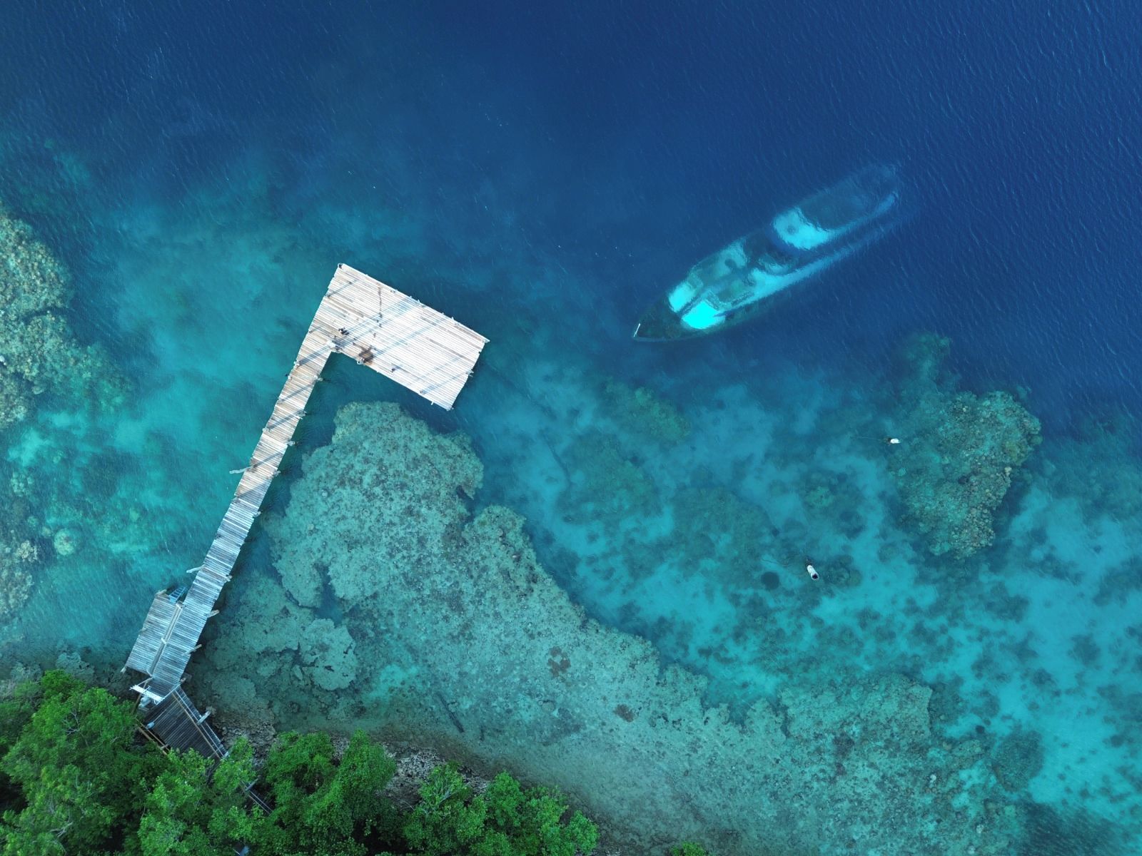 Ein Luftbild von tiefblauem Meer mit einer hölzernen Anlegestelle, die ins Wasser führt, einem in der Nähe vor Anker liegenden Boot und einer kleinen Insel mit dichter grüner Vegetation.