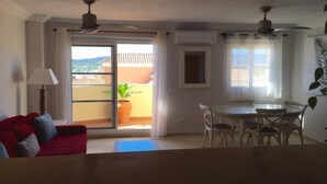 Une pièce avec un canapé rouge, une table à manger avec des chaises, un ventilateur de plafond et une porte-fenêtre coulissante donnant sur un balcon agrémenté d’une plante.