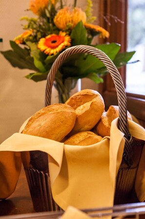 Una cesta de barras de pan con asa tejida, sobre una mesa con un florero de flores al fondo.