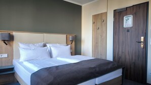 A hotel room with a bed, bedside lamps, a wooden door, and a wall-mounted information board. - St.-Michaels-Heim Hotel (Berlin)