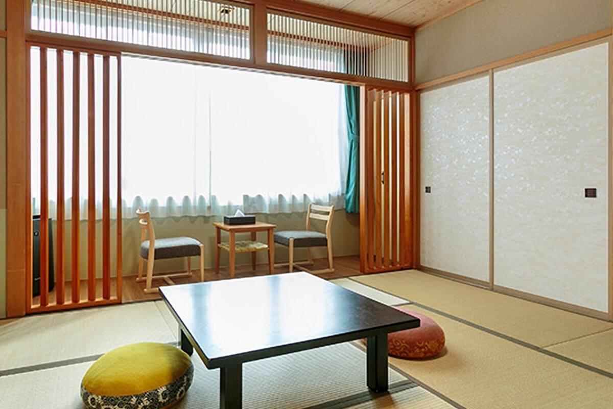 A traditional Japanese room with tatami flooring, a low table, two chairs, a sliding door, and a view of the outside.