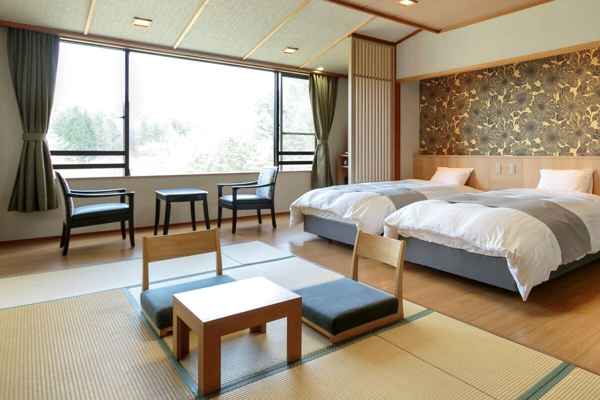 A hotel room with a large bed, a desk, two chairs, and a view of trees through the window.
