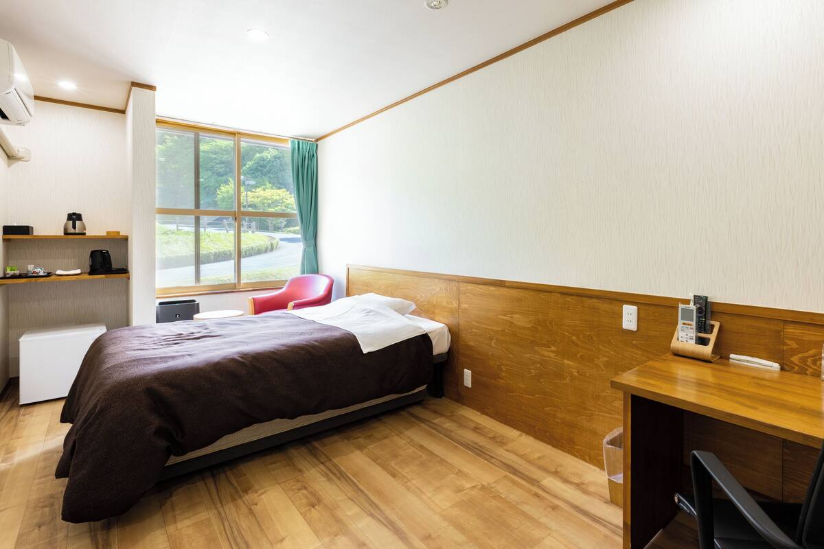 A hotel room with a bed, a desk, a chair, a window with curtains, a small table, and a shelf with a kettle and cups.