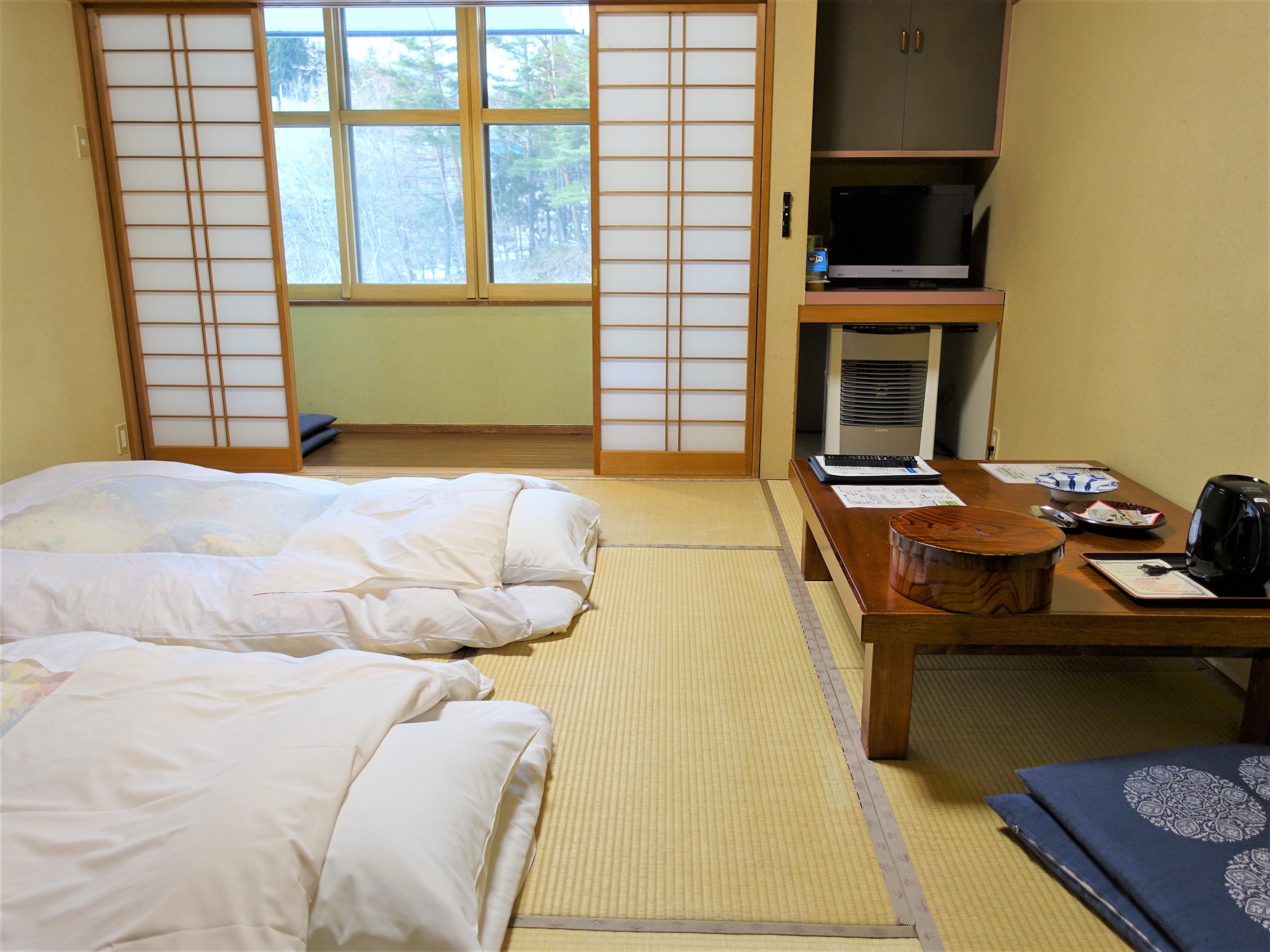 A traditional Japanese-style room with tatami flooring, a futon bed, a wooden table with a kettle, and a television on a cabinet.