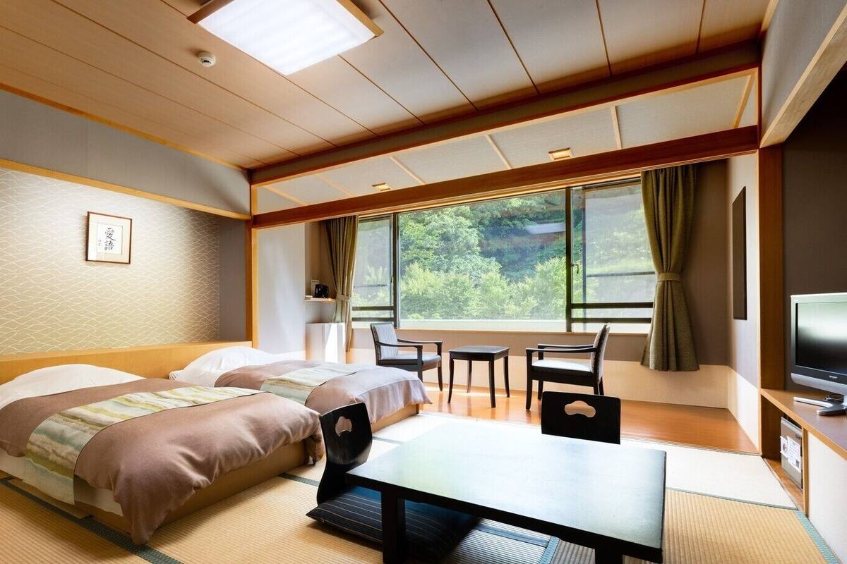 A hotel room with a bed, a desk, a chair, a television, and a view of greenery through the window.