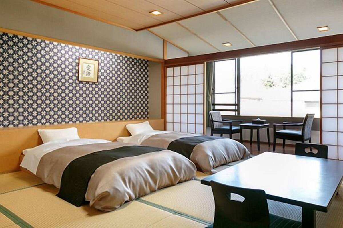 A traditional Japanese-style room with tatami flooring, two beds, a table, chairs, and a view through a shoji screen.