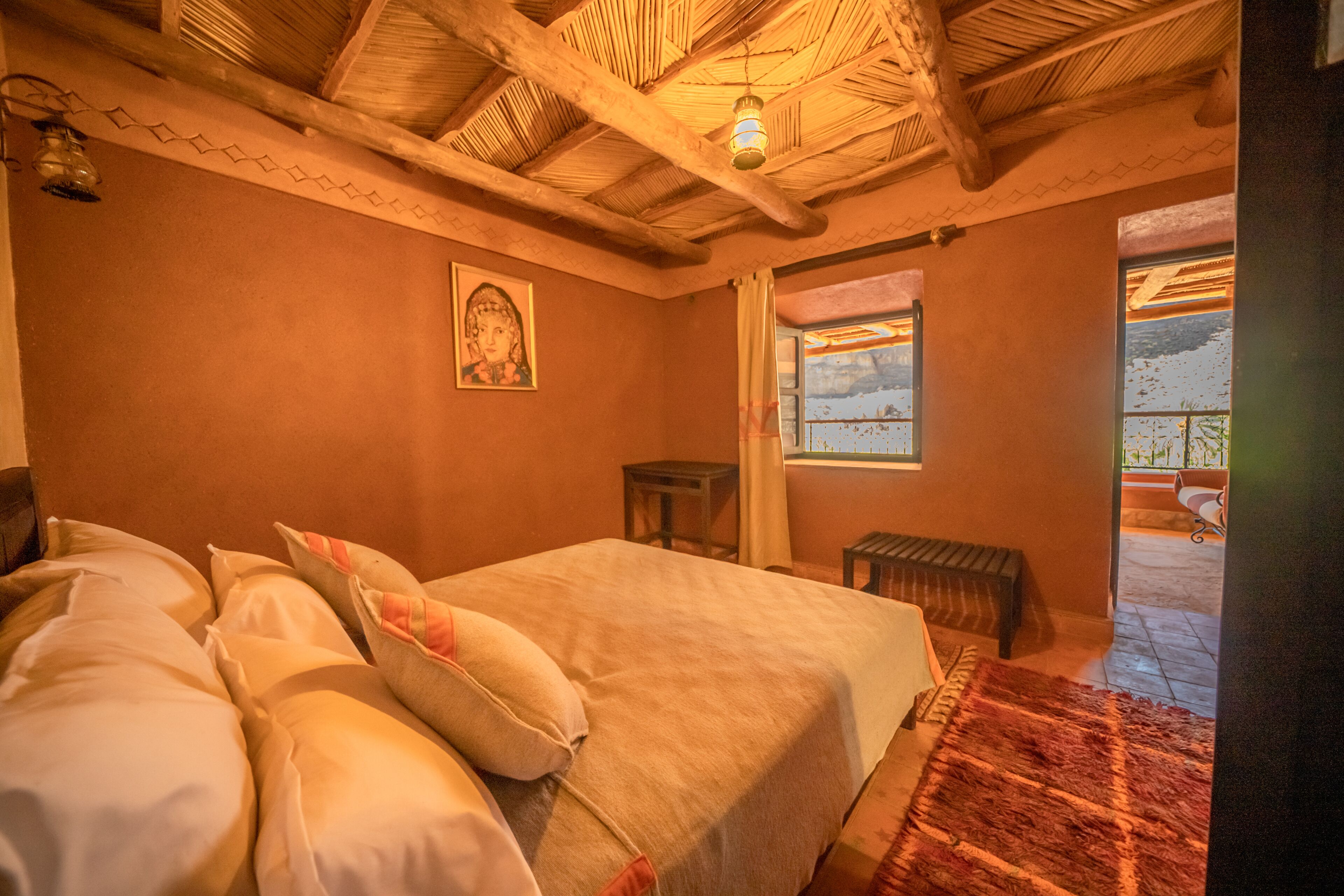 A bedroom with a bed, pillows, a wooden ceiling, a small table, and a window.