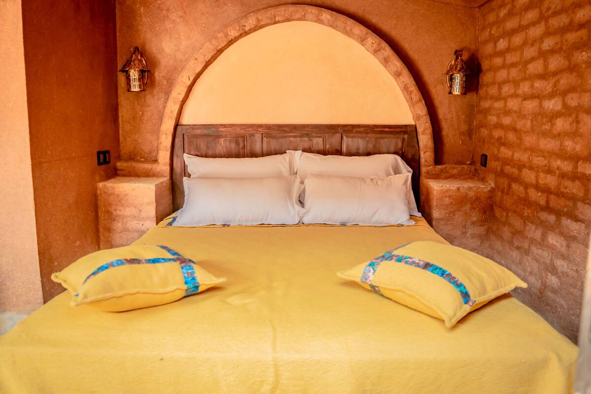 Una cama con cabecera de madera, dos almohadas y dos faroles fijados a la pared.