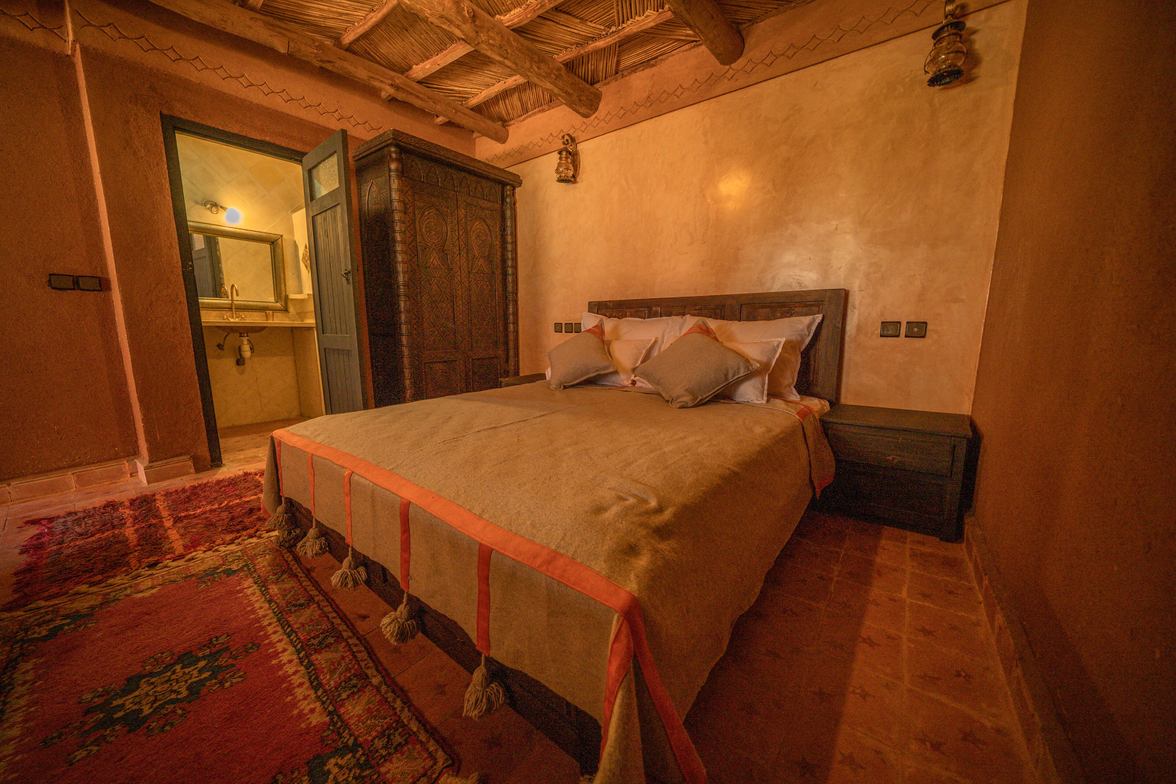 Une chambre à coucher avec un grand lit, une armoire en bois et une salle de bain visible à travers une porte ouverte.