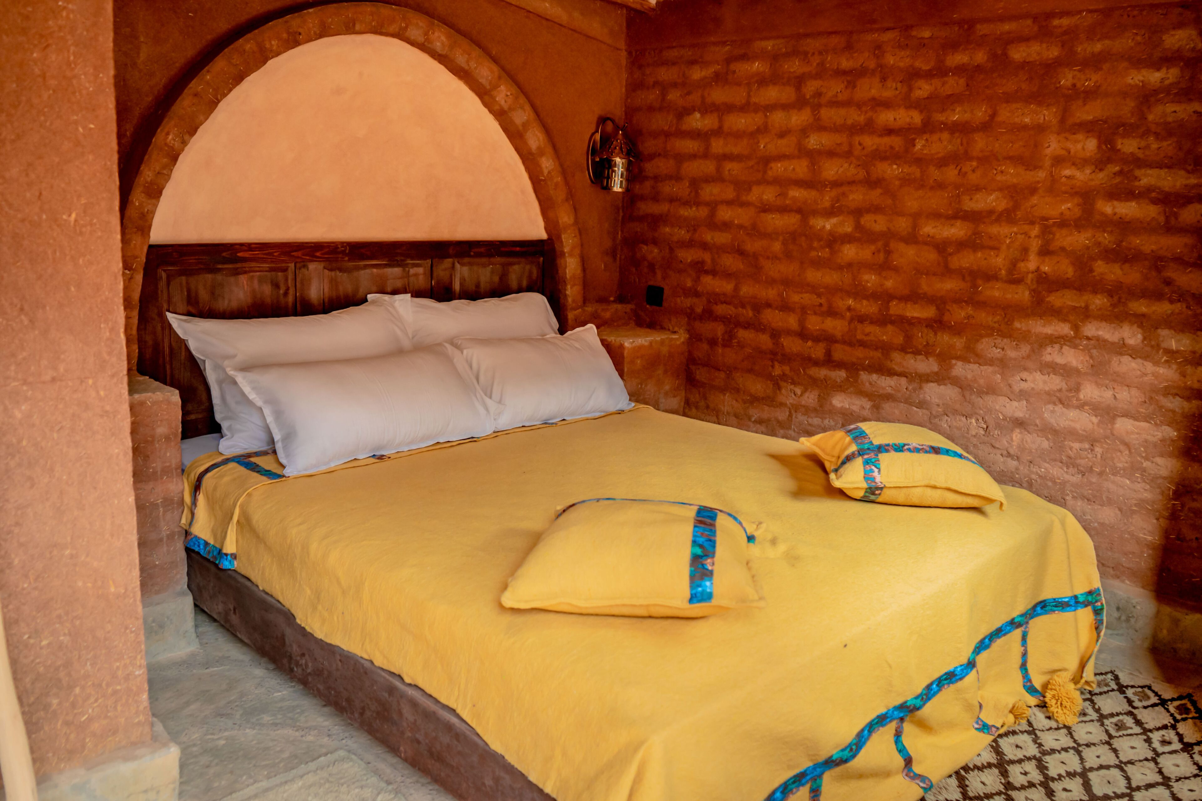 A bed with a yellow bedspread and pillows, a wooden headboard, and a brick wall.