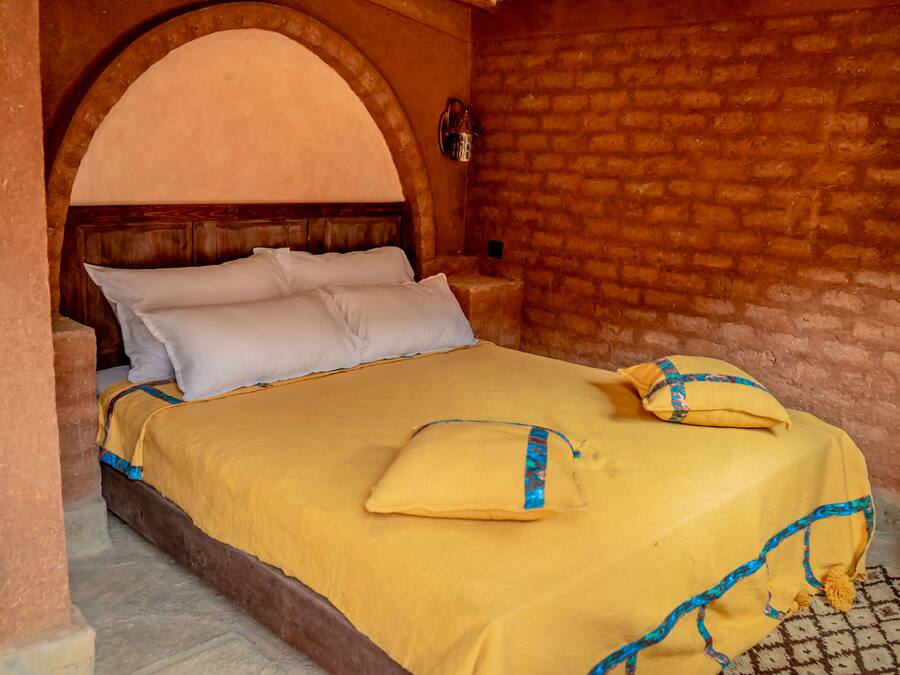 A bed with a yellow bedspread and pillows, a wooden headboard, and a brick wall.