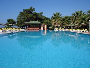 Une grande piscine extérieure, dotée de nombreux chaises longues et parasols, entourée d’arbres et agrémentée d’un espace de repos abrité.